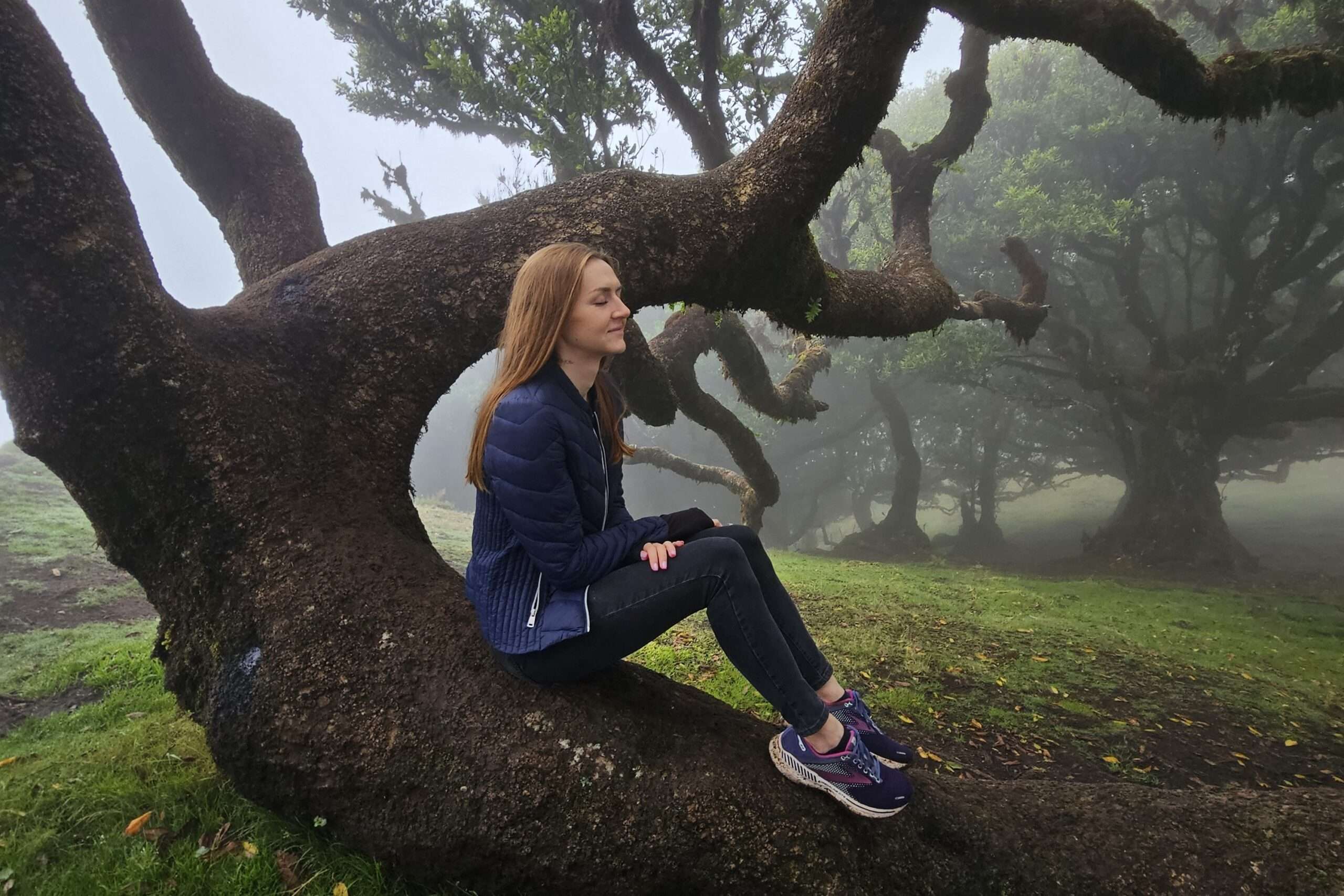 Kobieta siedząca na gałęzi drzewa w lesie, w spokojnej, zamglonej scenerii – ujęcie symbolizujące inspirację naturą i wewnętrzne wyciszenie, które sprzyja praktyce, jaką jest malarstwo intuicyjne. ( Intuitive painting)